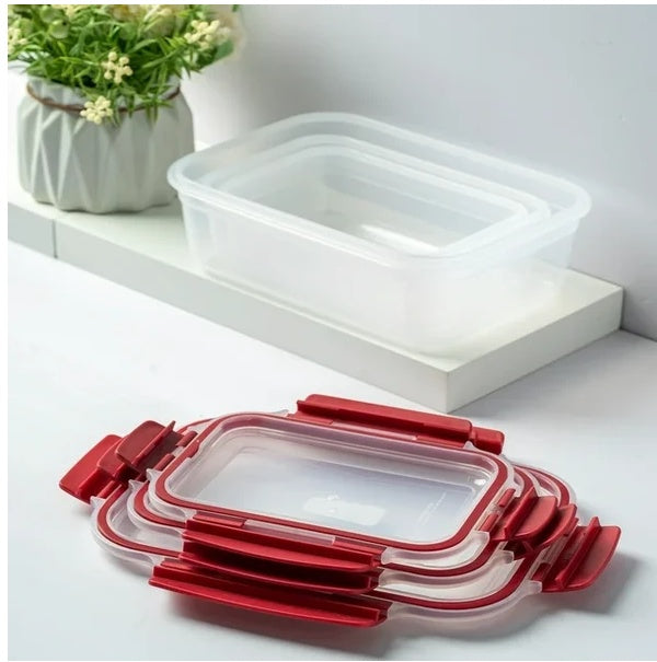 Set of clear food containers with red lids on a white surface, with a plant in the background.