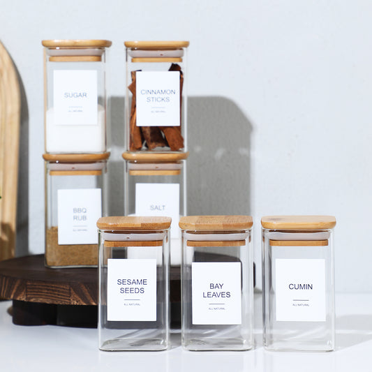 Set of glass jars with wooden lids labeled with various spices on a white background