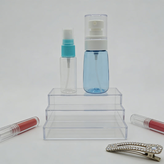 Two clear spray bottles with blue and turquoise caps on a white background, with lip gloss and a hair clip in the foreground.